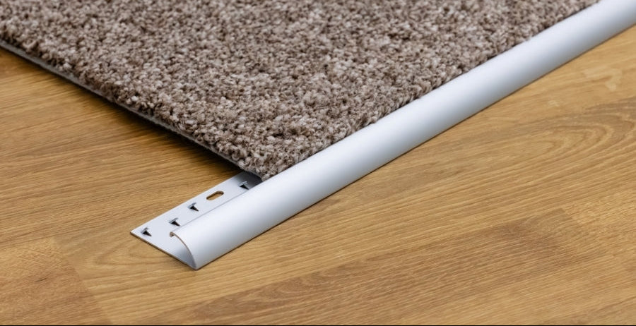 A close-up of a carpet transition strip in white, shown at the corner where a brown textured carpet meets a light wood floor. The logo 'MW Profiles' is visible in the top left corner.